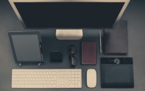 A top down image of an organized workspace including a computer monitor, keyboard, mouse, planner, glasses, pen, phone and charger, journal and electronic drawing tablet.