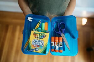 A child holds open a plastic pencil case fulled with glue sticks, colored pencils and other school supplies.