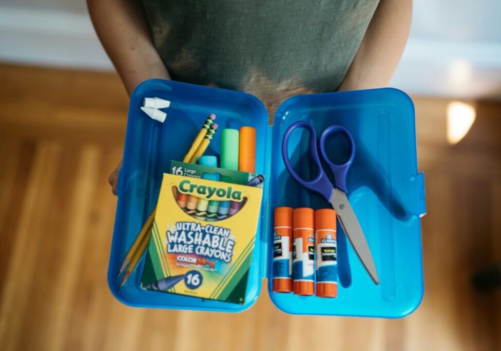 A child holds open a plastic pencil case fulled with glue sticks, colored pencils and other school supplies.