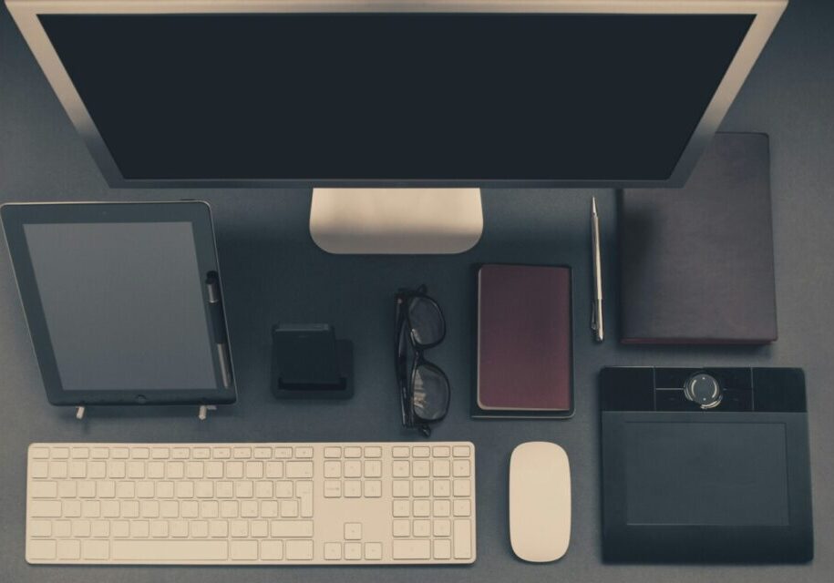 A top down image of an organized workspace including a computer monitor, keyboard, mouse, planner, glasses, pen, phone and charger, journal and electronic drawing tablet.